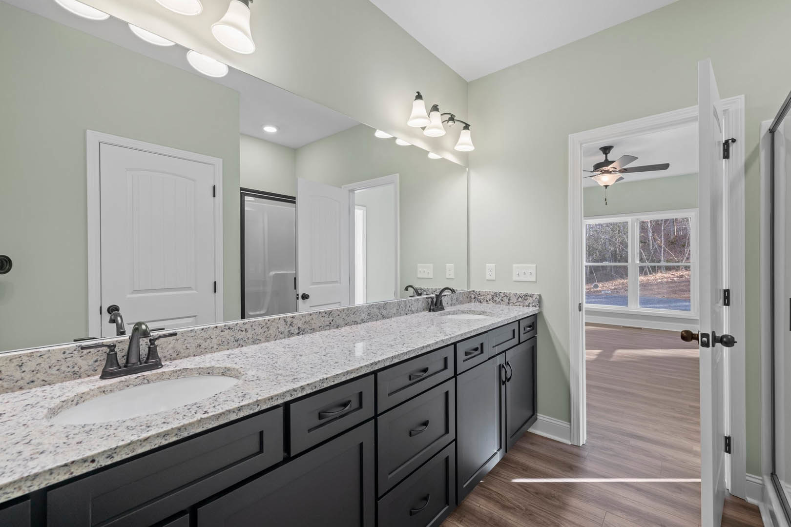 Bathroom with double white vanity, black hardware, large wall mirror, quartz countertop, white shower door with black handle, window overlooking trees, and modern light fixture.