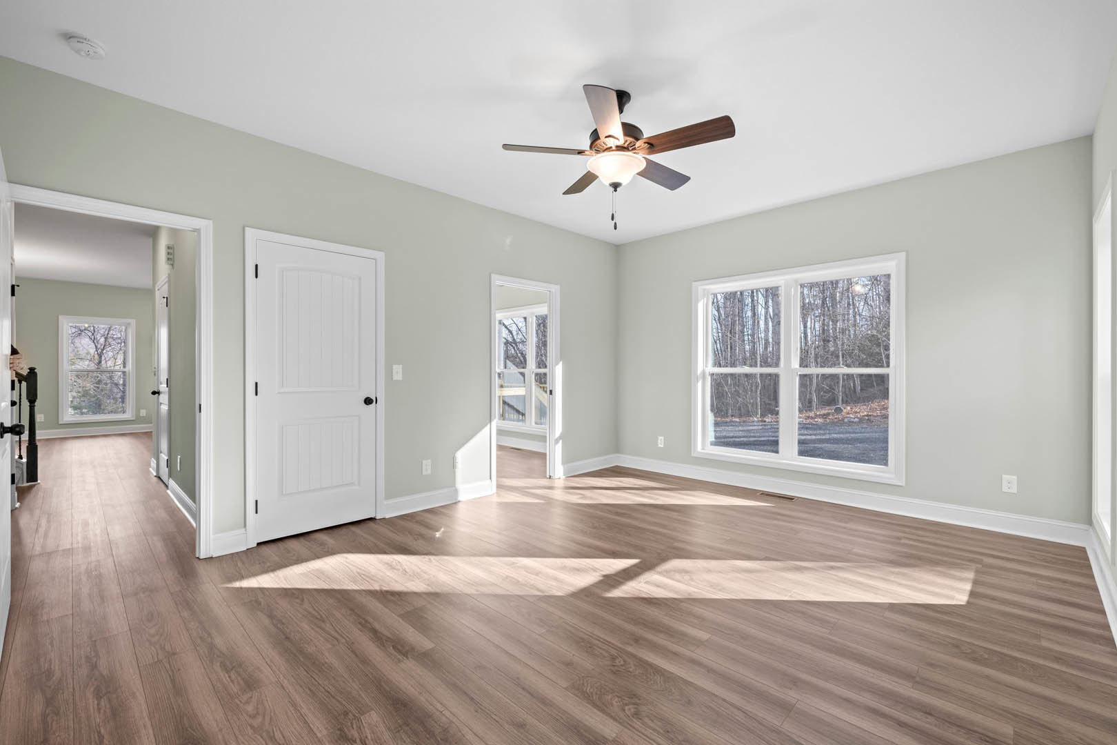 Ceiling fan with light fixture above wood flooring, white walls, white door with black knobs, large windows showing trees outside