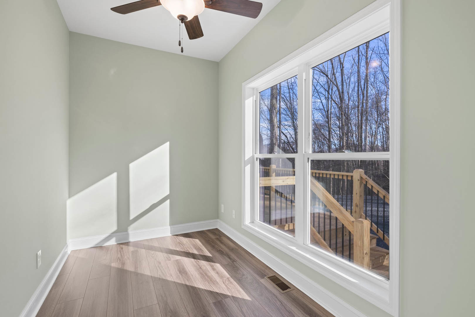 Wooden floor with white trim, ceiling fan with light fixture, large window overlooking trees, neutral walls, natural daylight filling the room