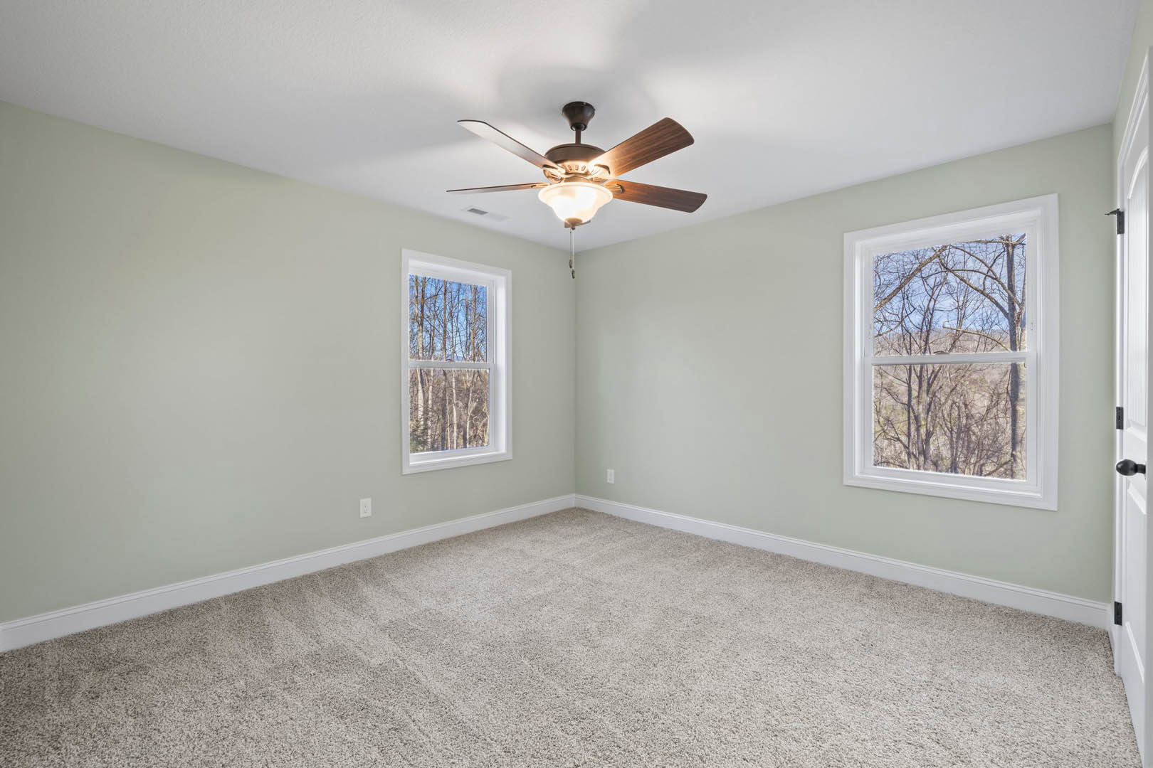 Neutral-toned carpeted room featuring a ceiling fan with integrated light, multiple windows framed by white molding, and views of green trees outside