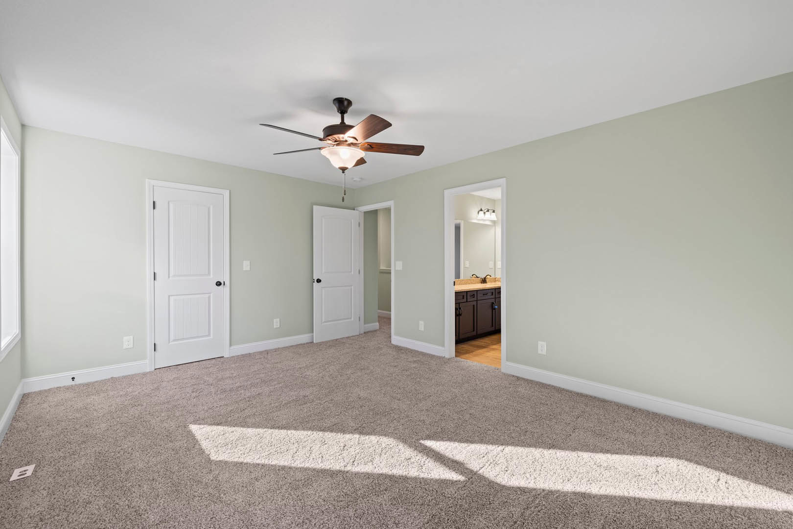 Carpeted bedroom with ceiling fan and light, white paneled door with black hardware, adjacent bathroom featuring a framed mirror, neutral walls and trim
