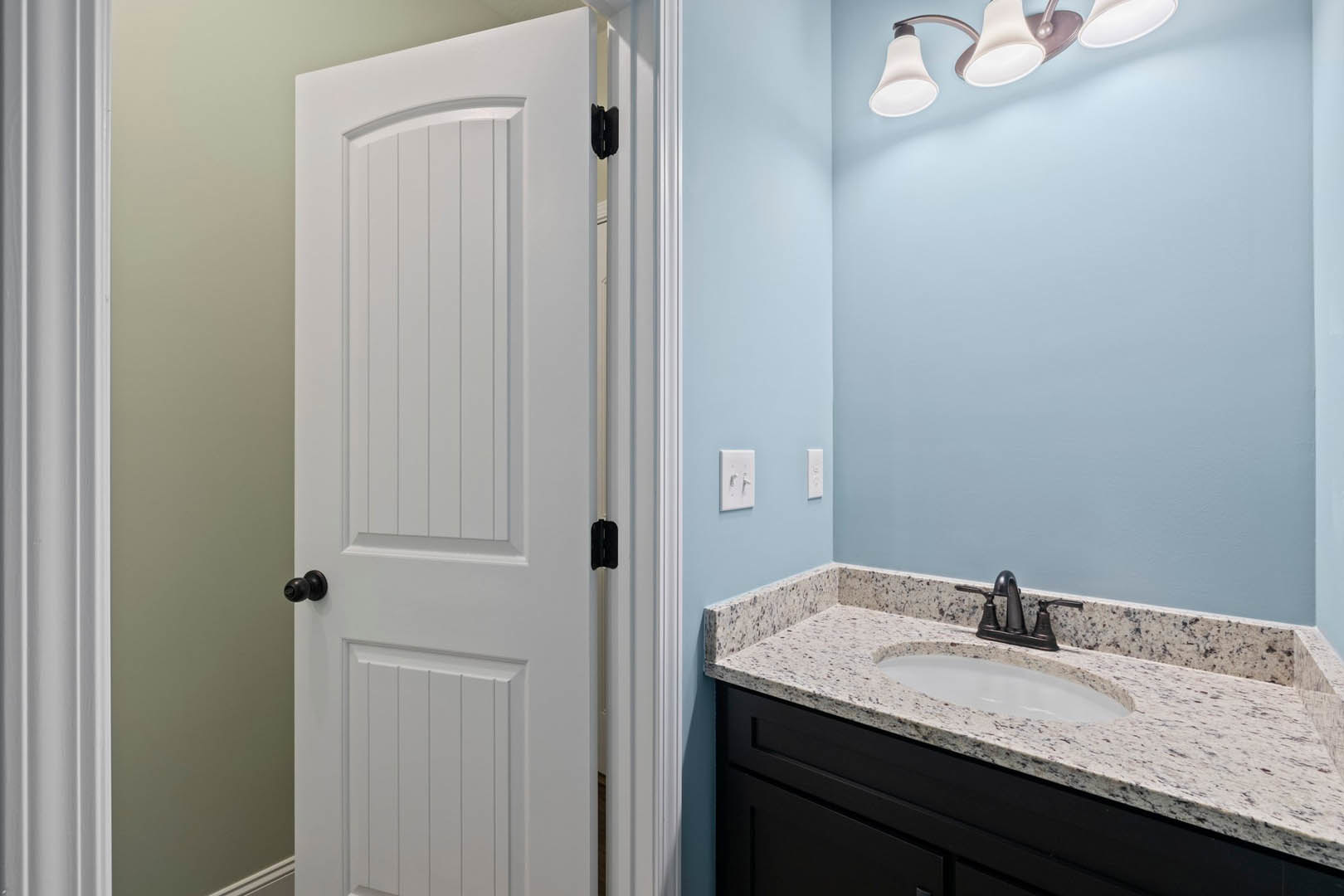 Bathroom with white paneled door, black door knob, white tile walls, built-in tub, wall-mounted sink, chrome faucet, rectangular mirror, and ceiling light fixture
