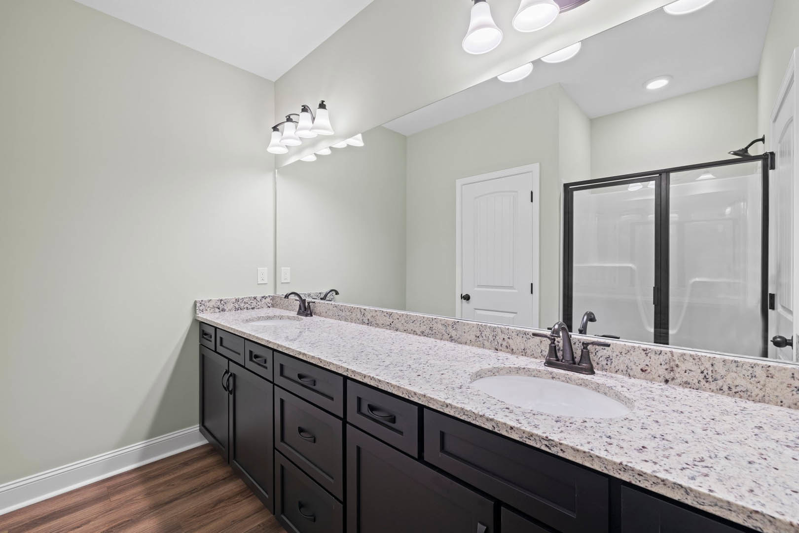 Bathroom with double white sinks, black faucets, large wall mirror, glass shower enclosure, white door with black hardware, tile flooring, and modern light fixture