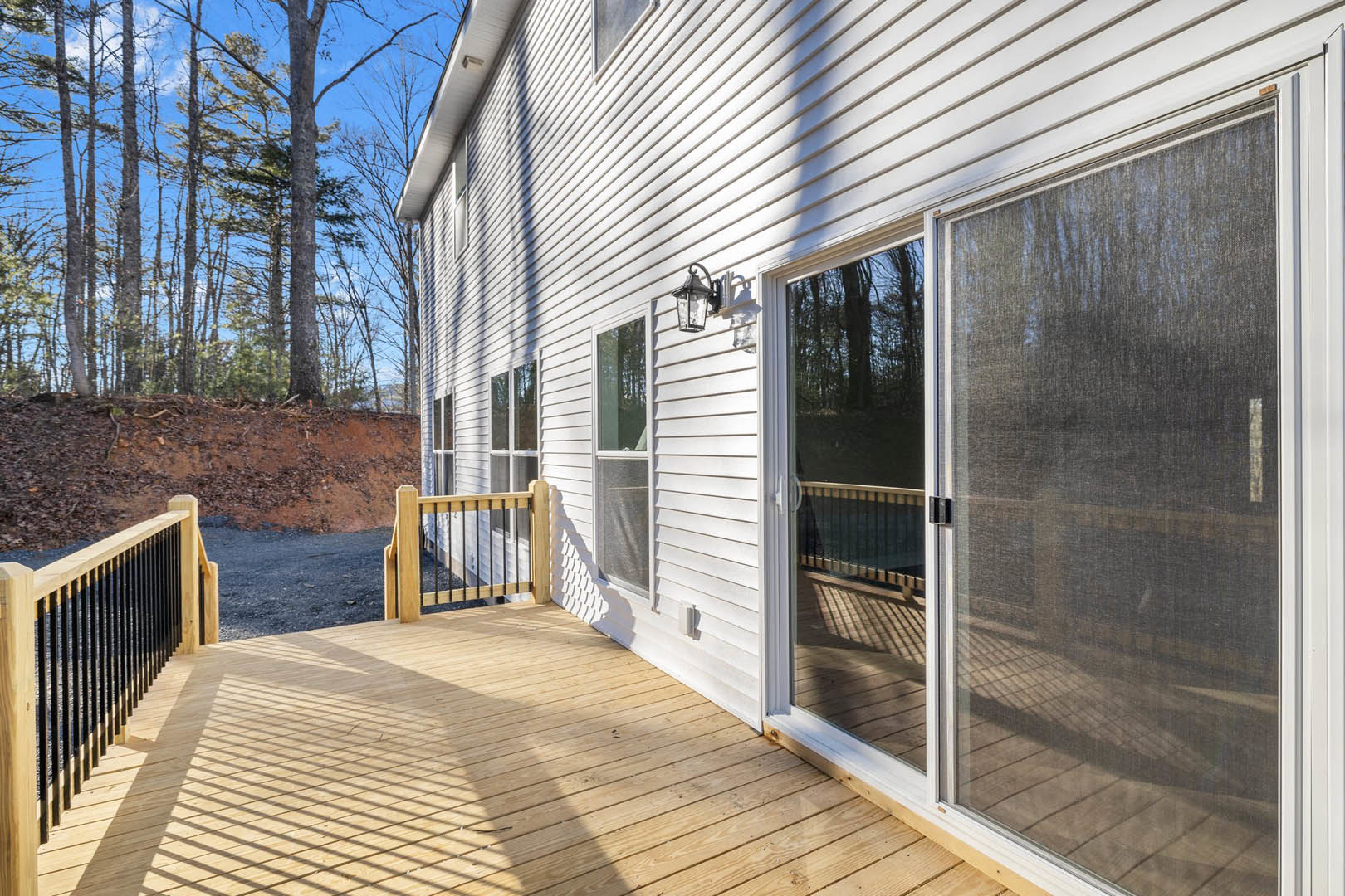 Wood deck with horizontal metal railing, sliding glass doors, screen door, exterior light fixture, surrounded by tall trees
