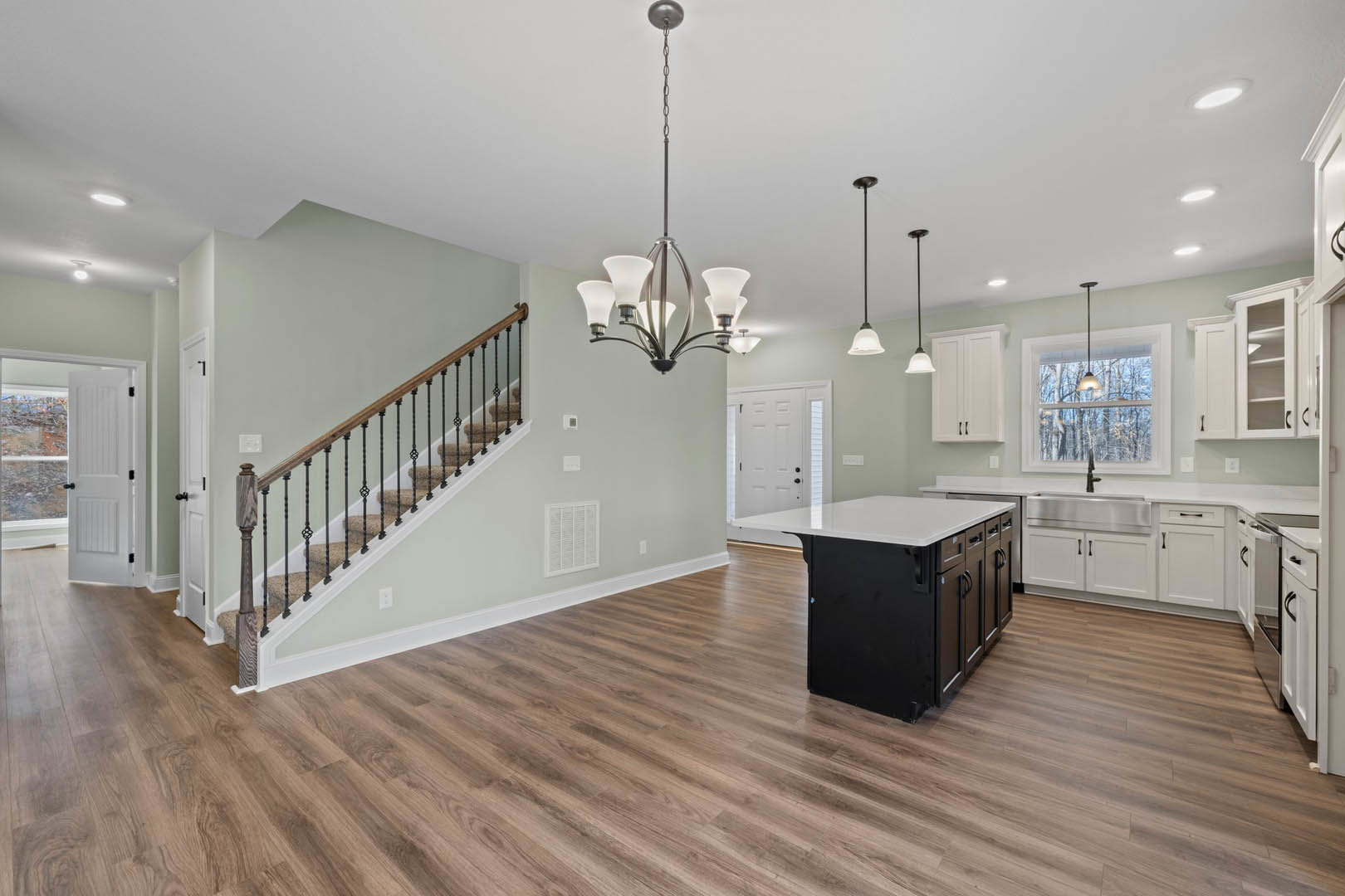 Open kitchen and dining area with wood flooring, white kitchen island, modern cabinetry, adjacent staircase with wood steps, chandelier with white shade, recessed ceiling lights
