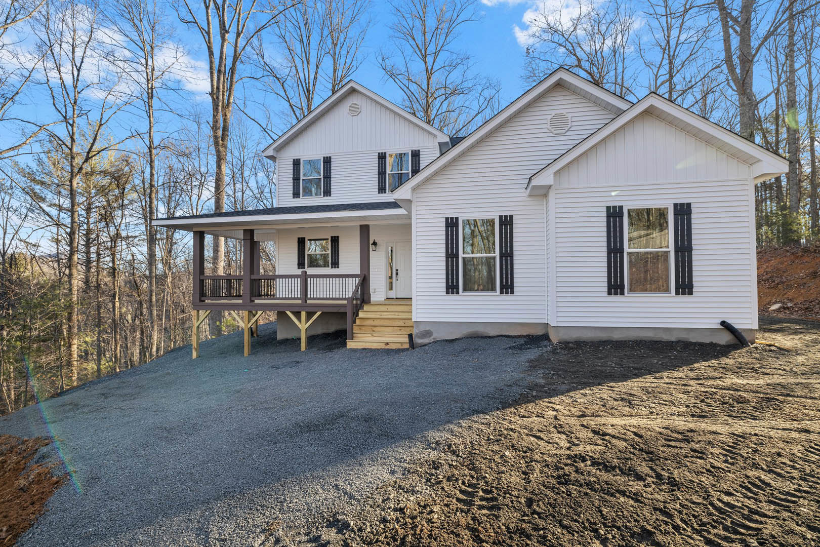 Two-story house with light siding, covered front porch with white railings, gravel driveway, exterior staircase, large windows reflecting trees, and landscaped yard.