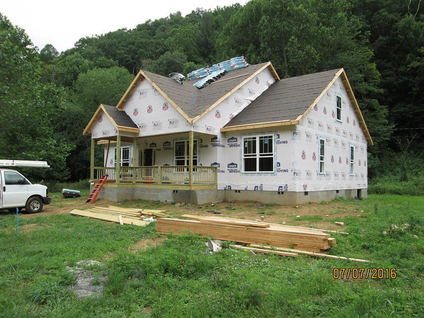 Wood-framed house under construction with exposed insulation on roof, white van parked nearby, pile of lumber on grassy yard, close-up of white door, yellow numbers on green grass.