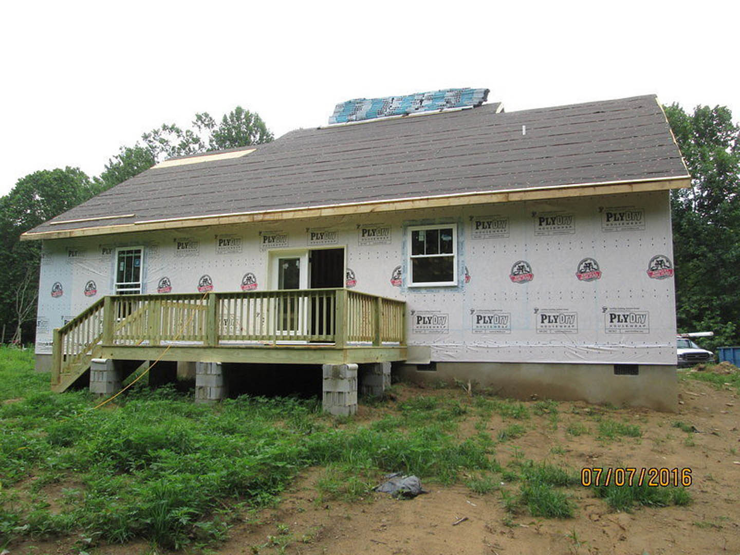 Partially built house with exposed insulation, wooden deck with railing, covered porch, white-framed window, roof topped with stacked plastic, and construction logo visible on