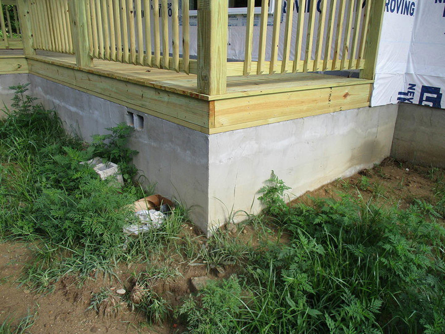 Front porch with wooden railing, stone wall base, green grass and weeds along the edge, blue letter sign on white siding