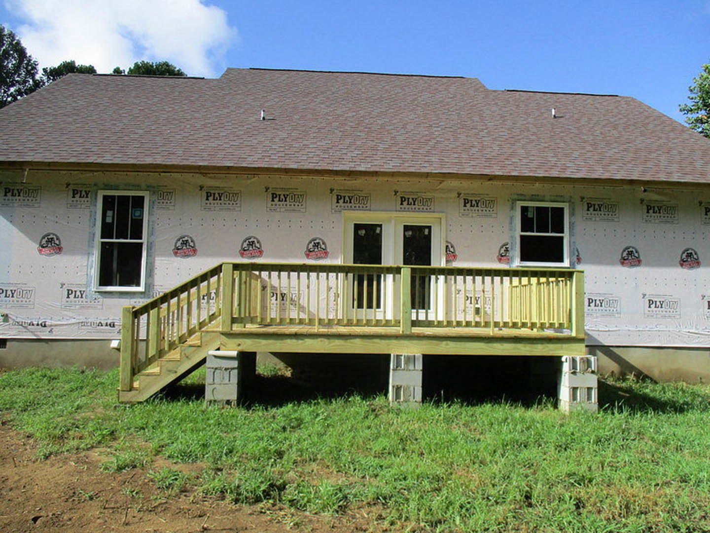 Two-story home with white-framed windows, wooden deck with railing, covered porch, white door, and grassy lawn under partly cloudy sky