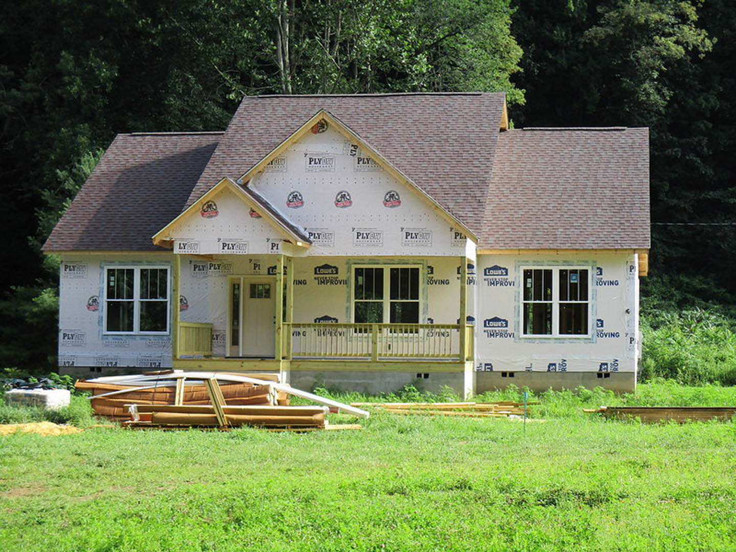 Wood-clad farmhouse exterior with covered porch, white door featuring window, yellow railing, and grassy yard with mature trees