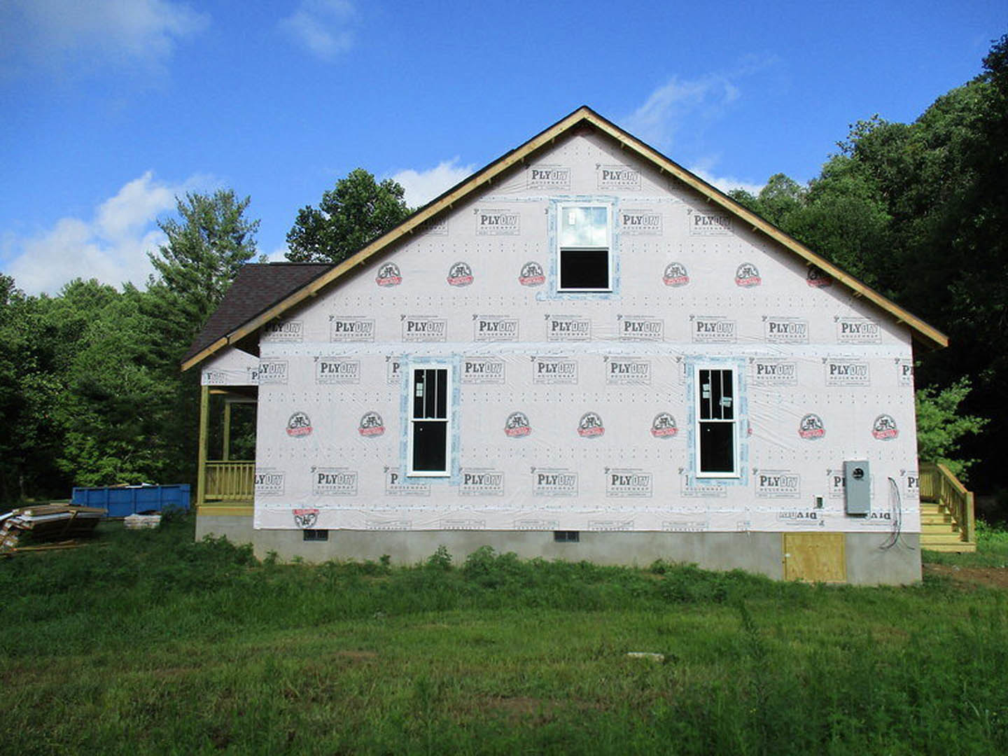 White exterior walls with large windows, grassy lawn in front, wooden staircase visible near entry, surrounded by trees and plants under a partly cloudy sky