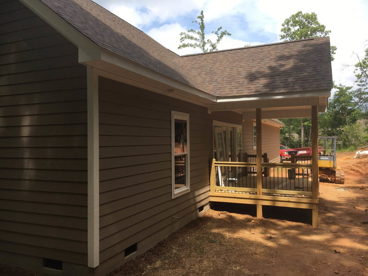 Two-story home with light siding, covered front porch featuring wooden railings, multiple windows reflecting sunlight, and a deck area surrounded by mature trees.