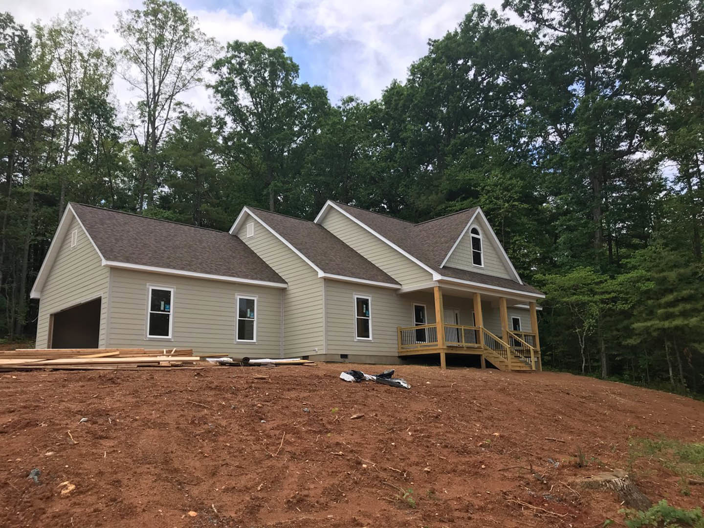 Partially built house with exposed wooden deck and railing, white-framed window, dirt yard scattered with construction debris, surrounded by tall trees under blue sky with clouds