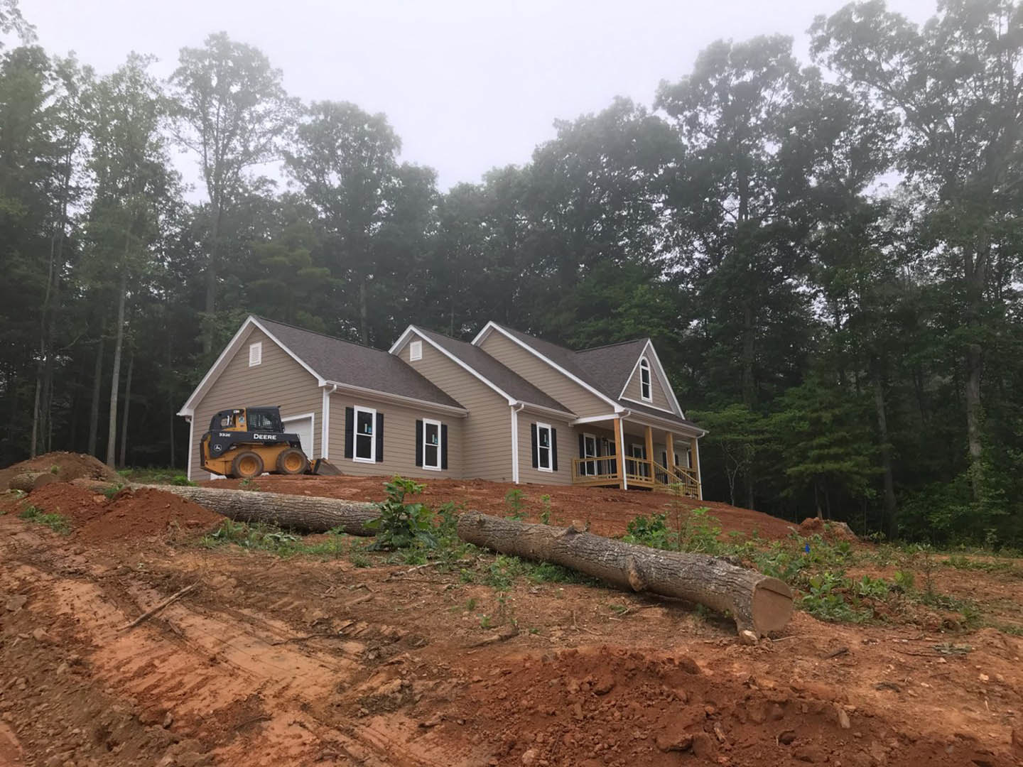 Modern cottage with white roof, large windows with white frames, tractor parked on soil in front, log lying nearby, group of trees in foggy background