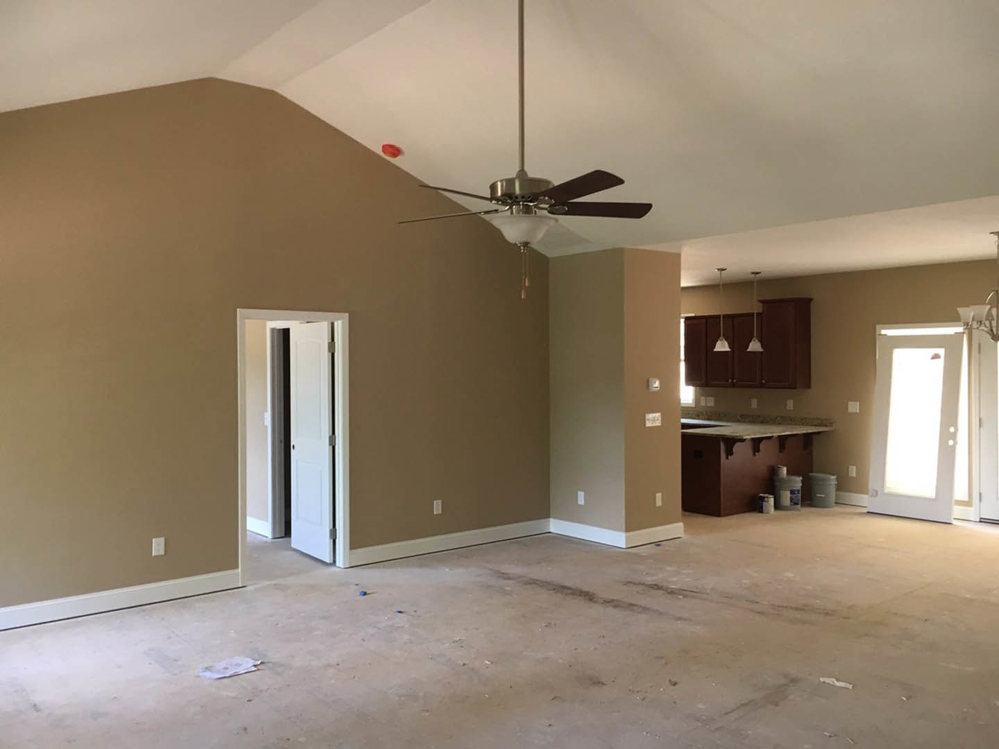Open-concept room with ceiling fan, white paneled door, pendant lights, kitchen with light countertops, paint bucket on counter, and unfinished flooring.