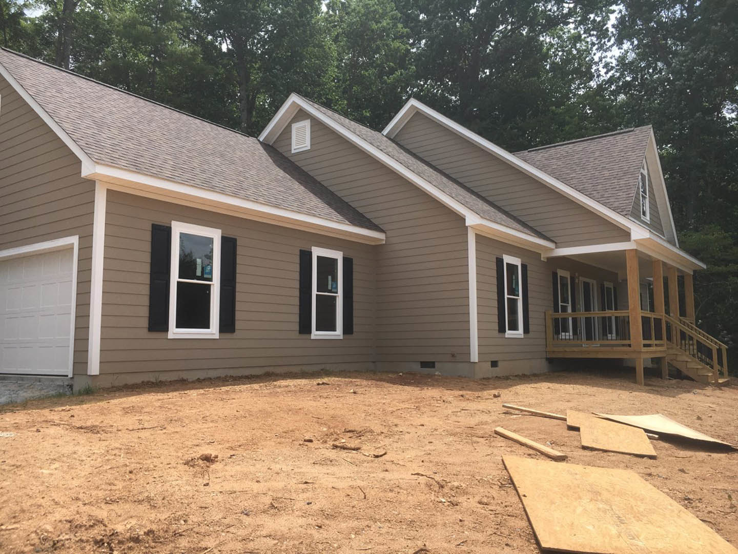 Two-story house under construction with white siding, several windows, dirt yard scattered with sticks and branches, surrounded by trees