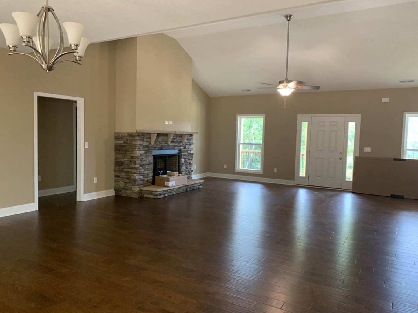 Living room with wide plank hardwood floors, stone fireplace with brick surround, white glass-paneled door, large window overlooking trees, and decorative chandelier.