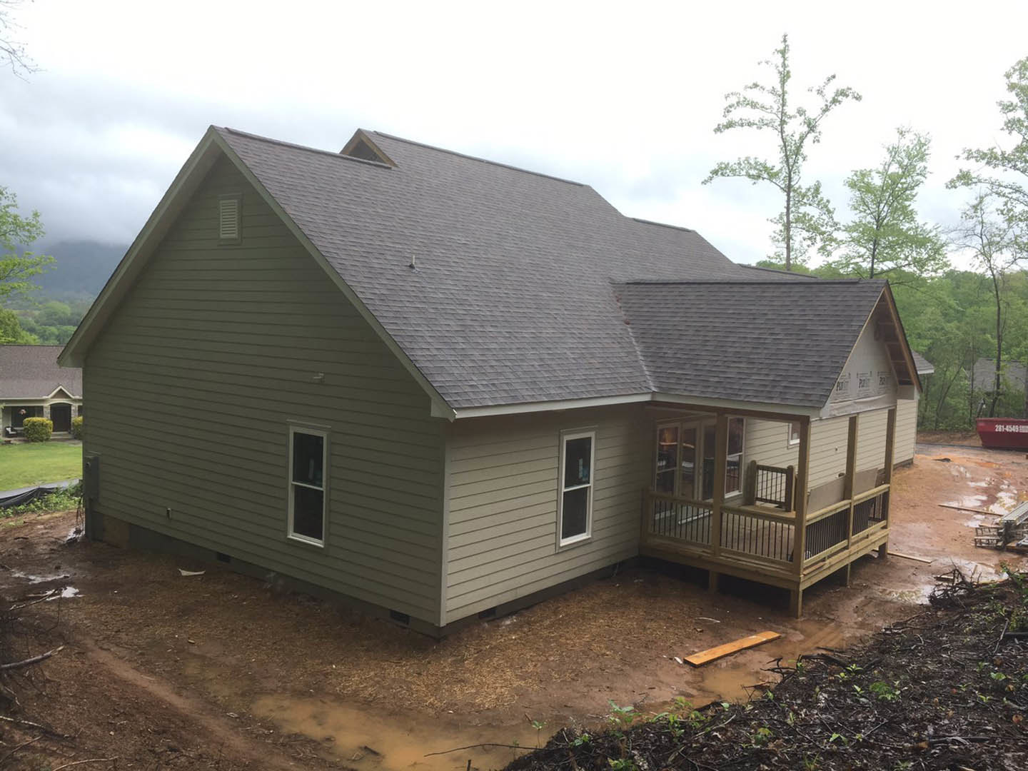 Two-story home with gray siding, white trim, covered front porch with wood decking, large windows, gabled roof, and landscaped yard with mature trees