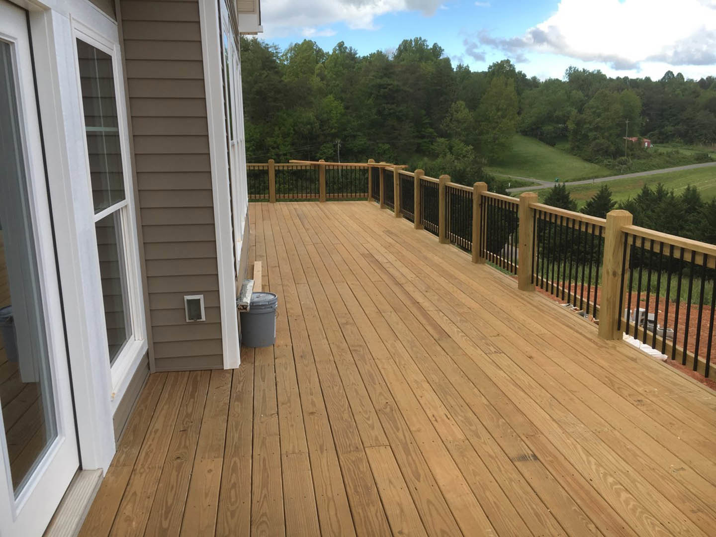 Wooden deck with railing, grey lidded bucket, window on exterior wall, trees and blue sky with clouds in background