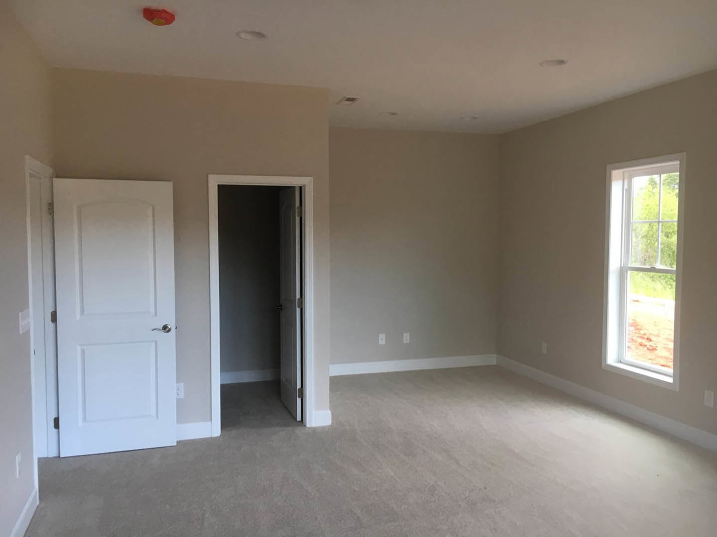 White carpeted room with open white door featuring silver handle, large window showing outdoor view, smooth plaster walls