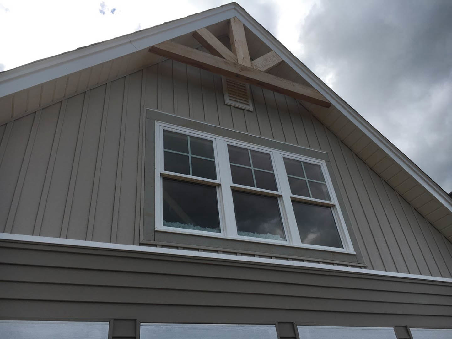 Modern home exterior with gray siding, black-framed windows reflecting cloudy sky, and dark shingle roof; rectangular vent visible above window.