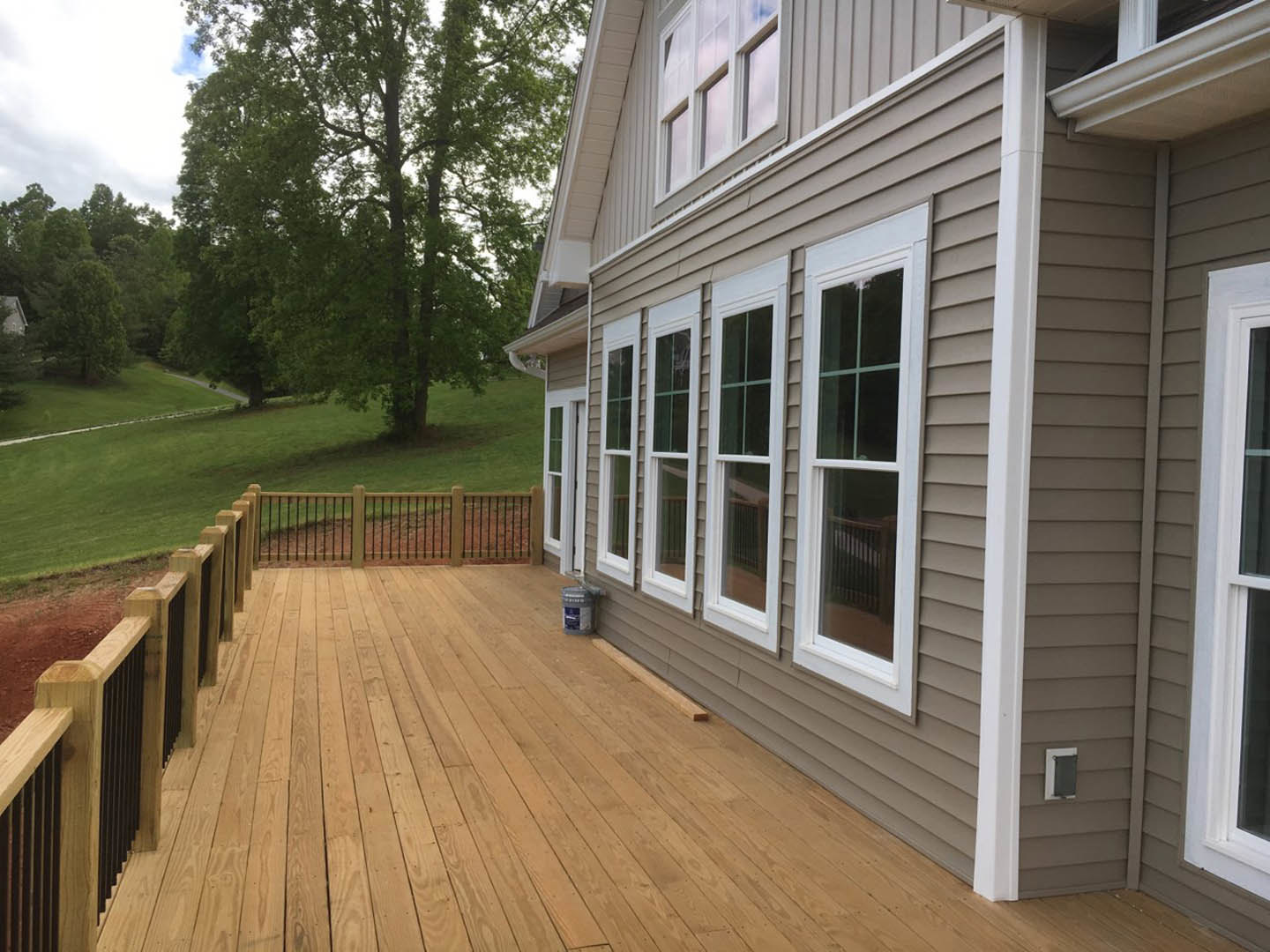 Modern home with light siding, wooden deck featuring a bucket of paint, lush green lawn, large tree, and partial view of fence and window.