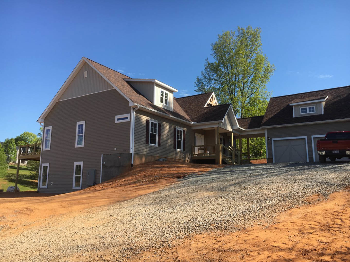 Two-story house with white siding, attached garage, red truck parked inside, gravel driveway, green-leafed tree in front yard, windows with dark trim, situated on a gentle hill.
