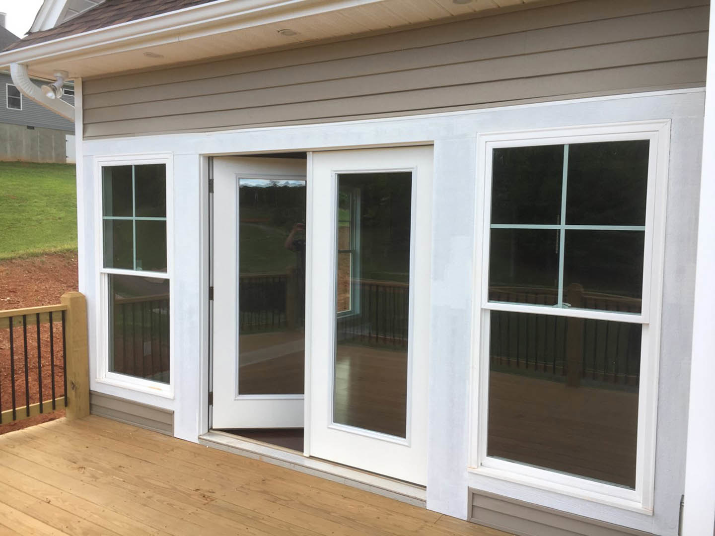 White double doors with glass panels opening onto a wooden deck, adjacent to a patio area, windows reflecting a nearby fence, and a wooden floor leading to the entry.