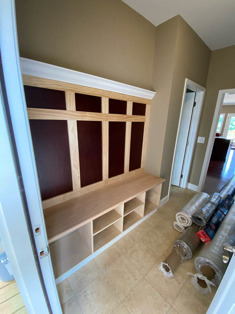 Wood bench beside rolled carpet rolls on hardwood floor, white walls, built-in shelving with wooden panel, door partially visible in custom home interior.