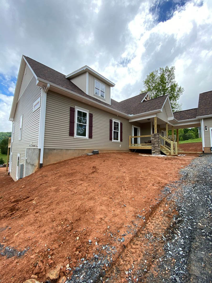 Partially built home with white-framed windows, stone pillar on porch, gravel-covered dirt hill in foreground, tree near roof, and unfinished deck.