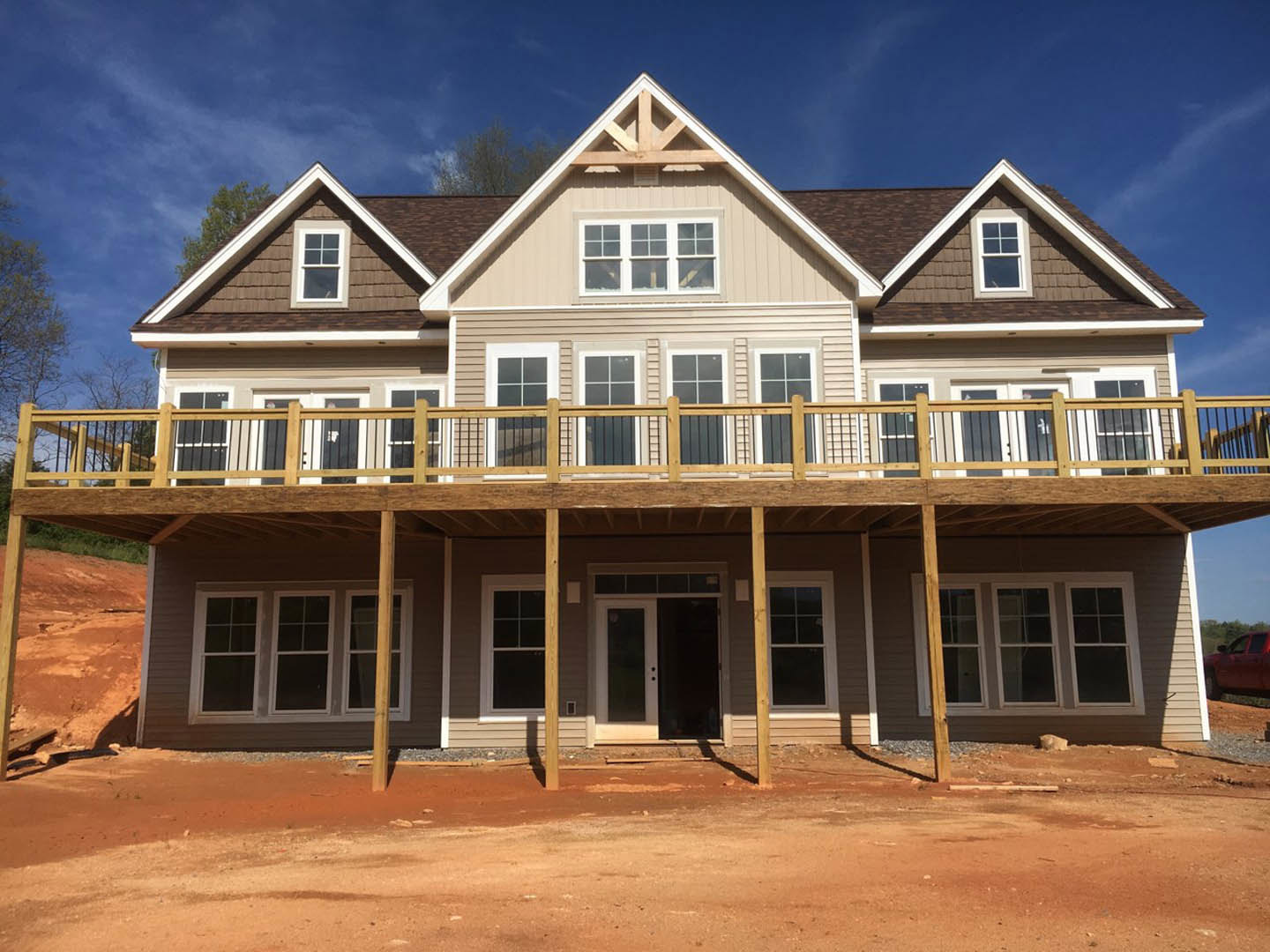 Wood-framed house under construction with exposed deck, white-framed windows, unfinished siding, dirt yard, and a red car parked nearby.