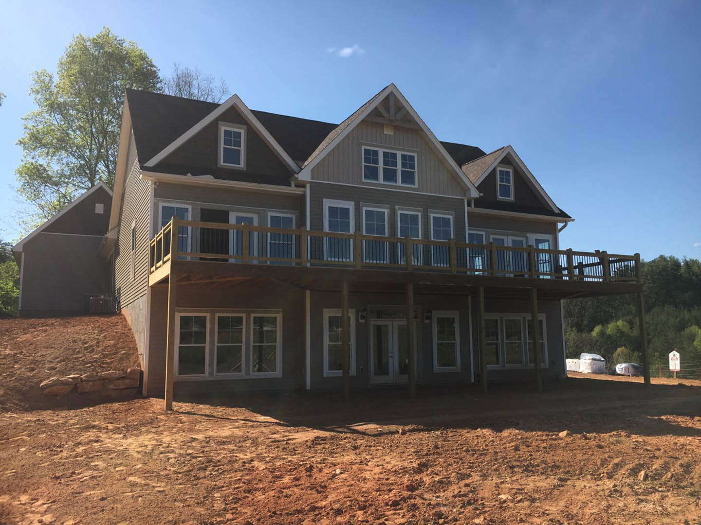 Two-story home with white-framed windows, wood deck with metal railing, surrounded by dirt field and mature trees, hillside visible in background under blue sky