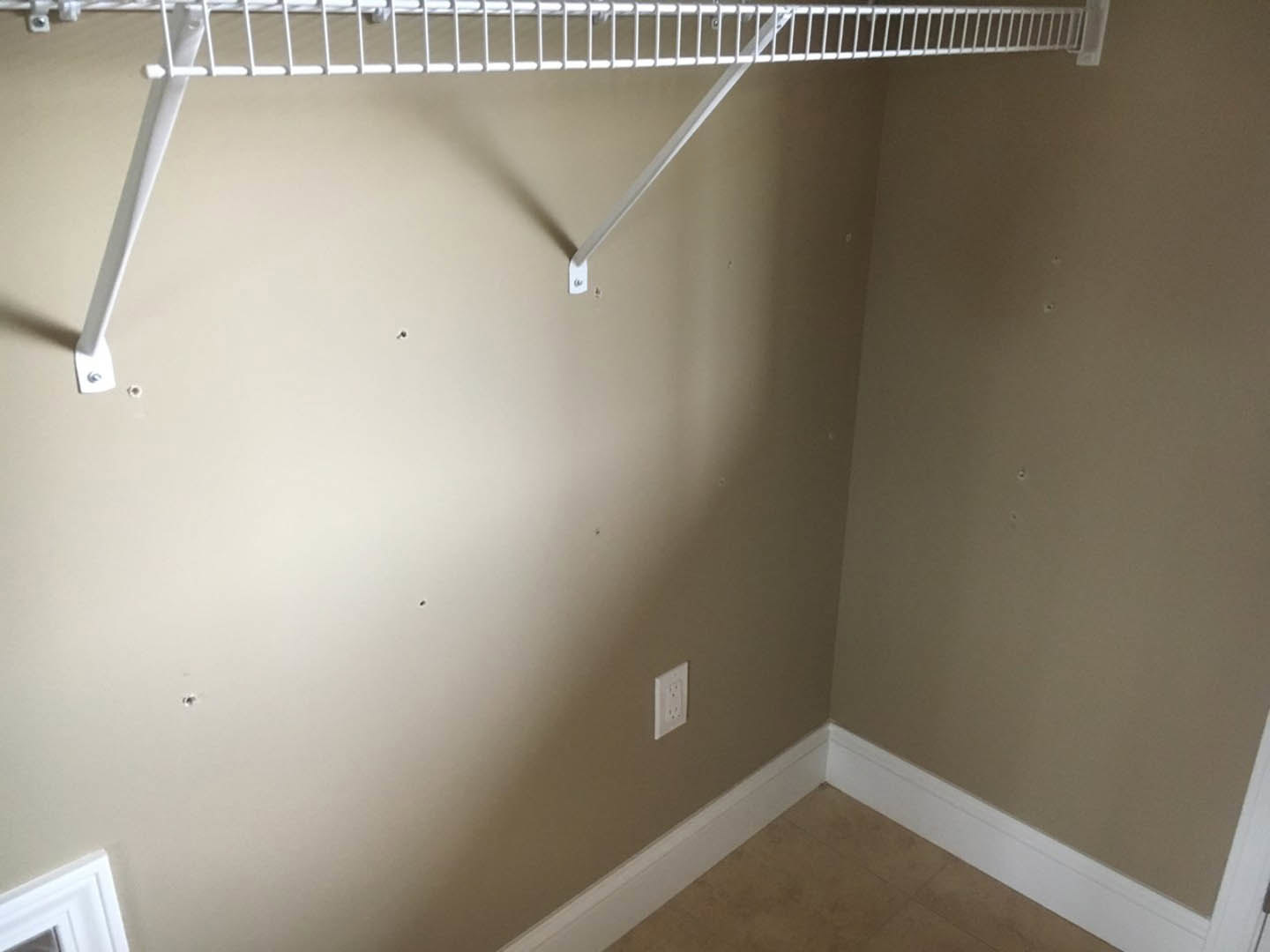 White metal shelf mounted on a plaster wall beside a white electrical outlet, with a close-up view of a white door and bar, in a room featuring a ceiling fan and light-colored