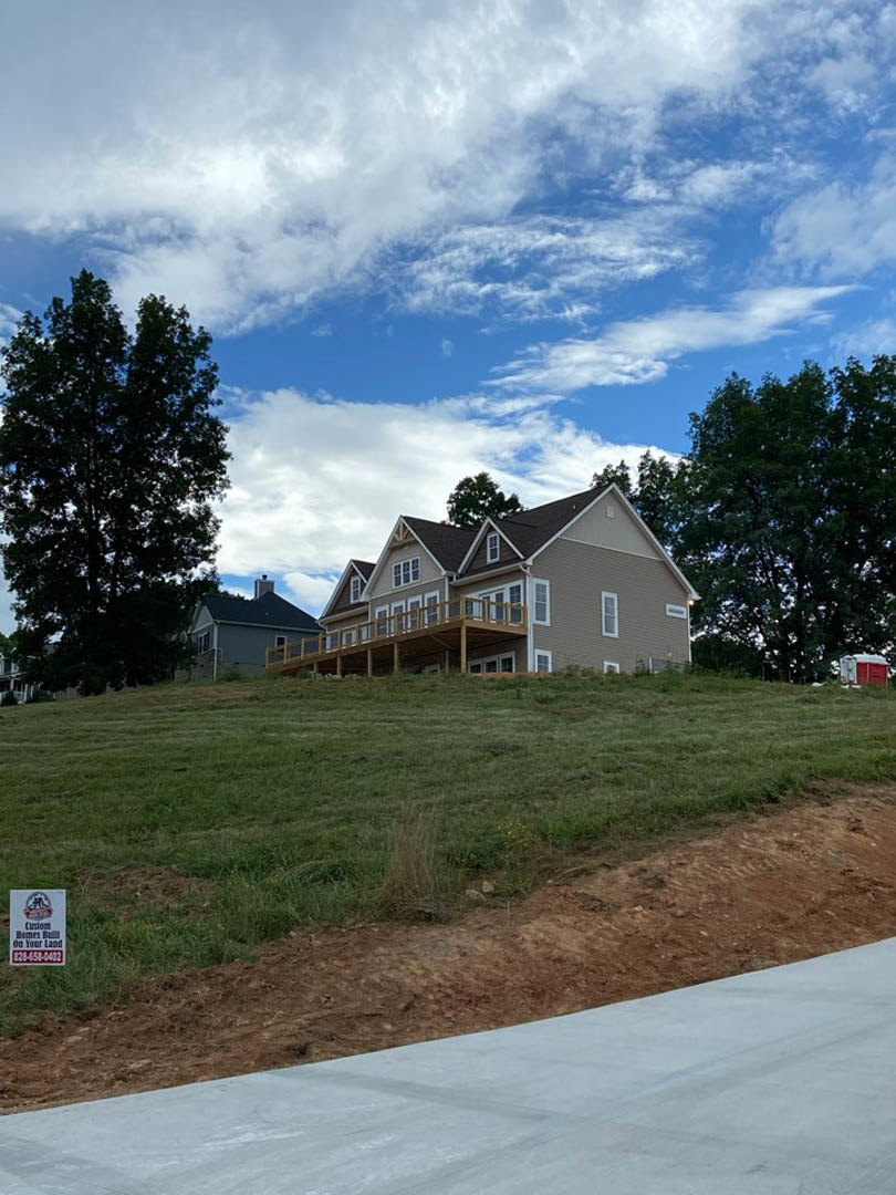 Modern cottage-style home with wood deck, large leafy tree, grassy hillside, dirt and concrete driveway, and a sign near the entrance under a partly cloudy sky