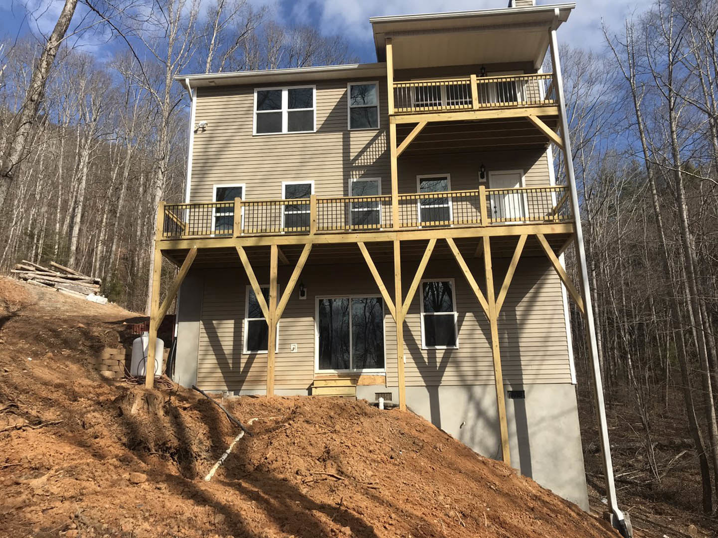 Two-story house with white siding, expansive wooden deck, large windows reflecting surrounding trees, hillside backdrop, and clear sky