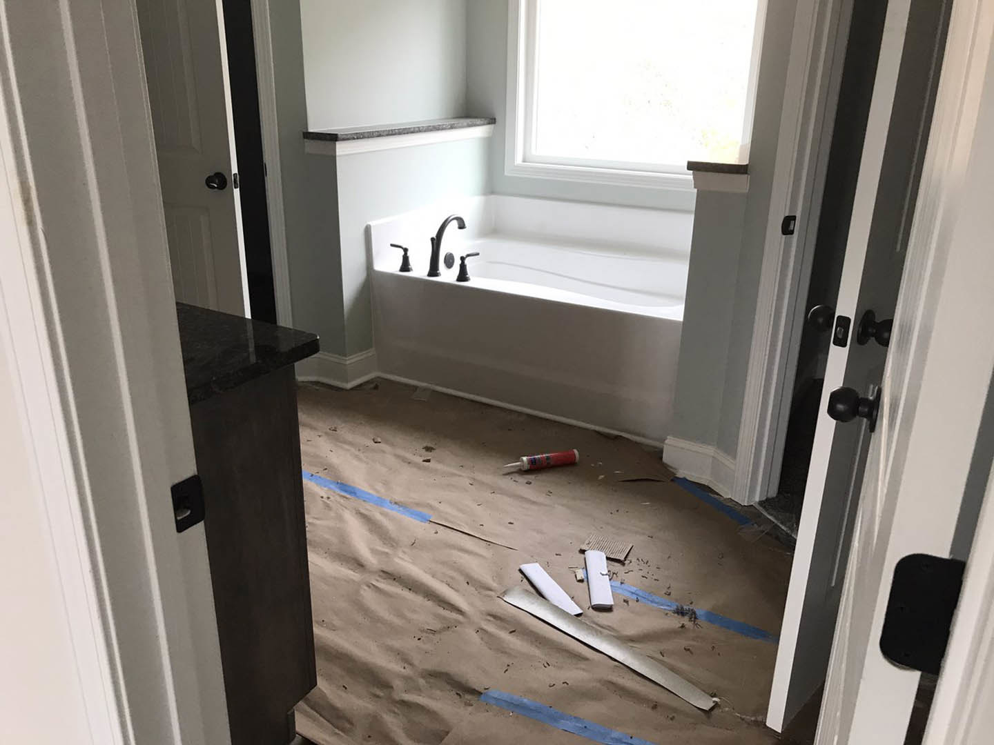 Freestanding white bathtub with chrome faucet, light-colored plaster walls, large window, wood flooring, and modern door in remodeled bathroom