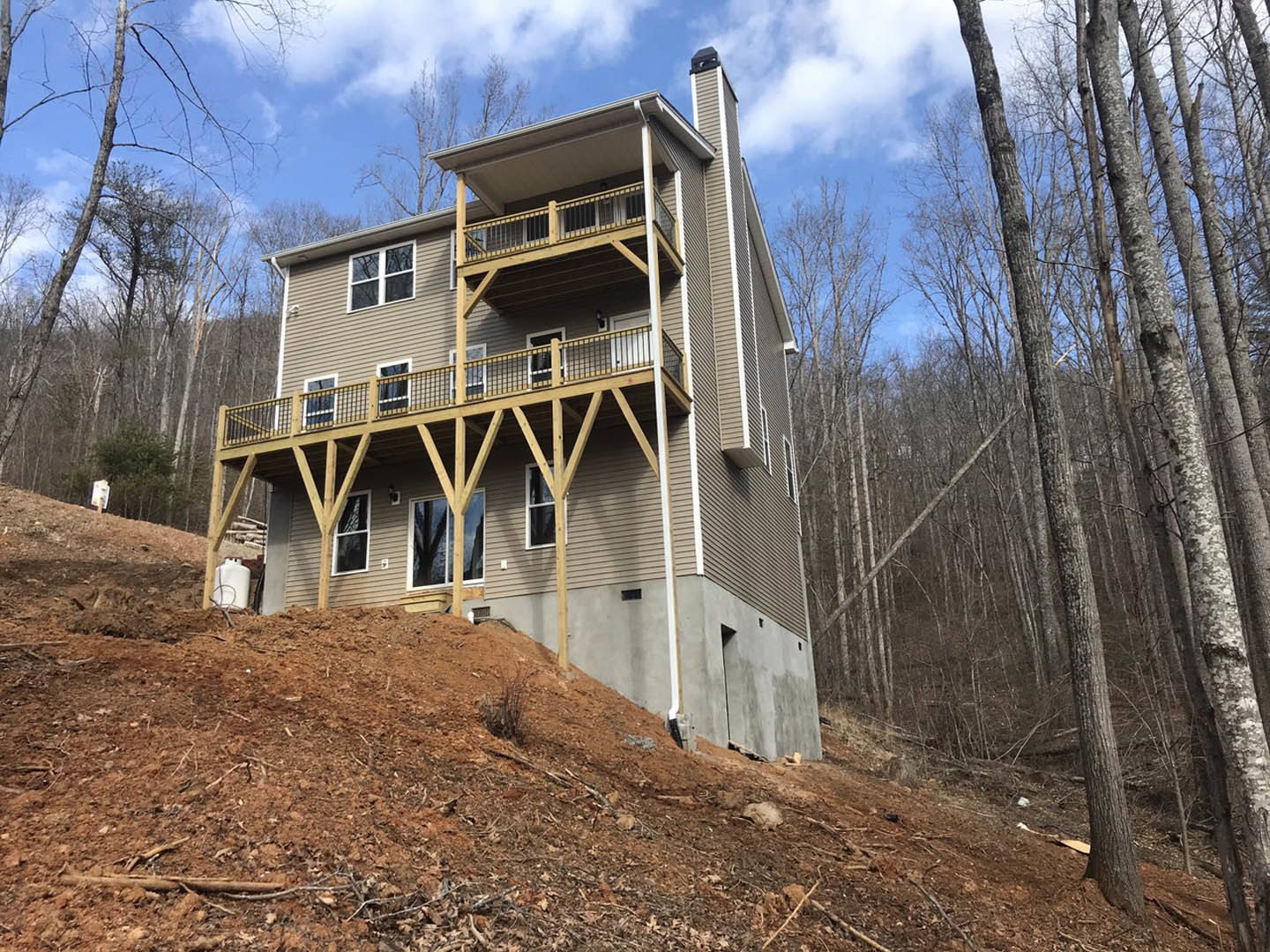 Two-story house with wood siding and large windows, situated on a grassy hill with mature trees and forest in the background