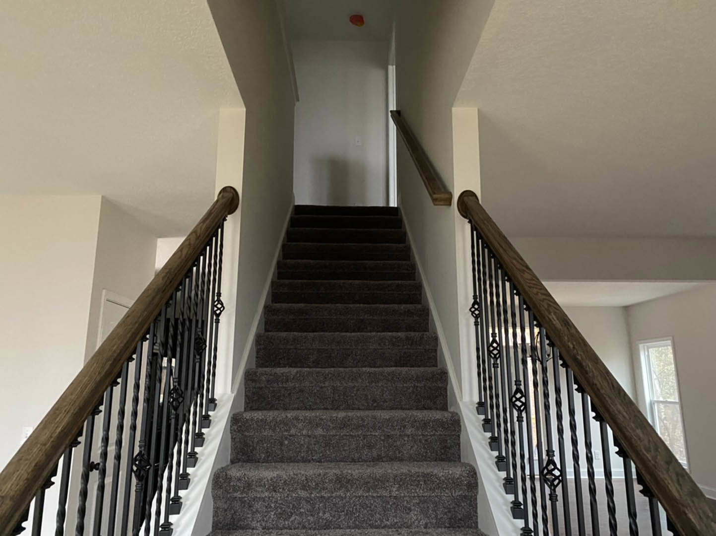 Staircase with grey carpet steps, wooden handrail, black balusters, and a window with a white frame on the landing