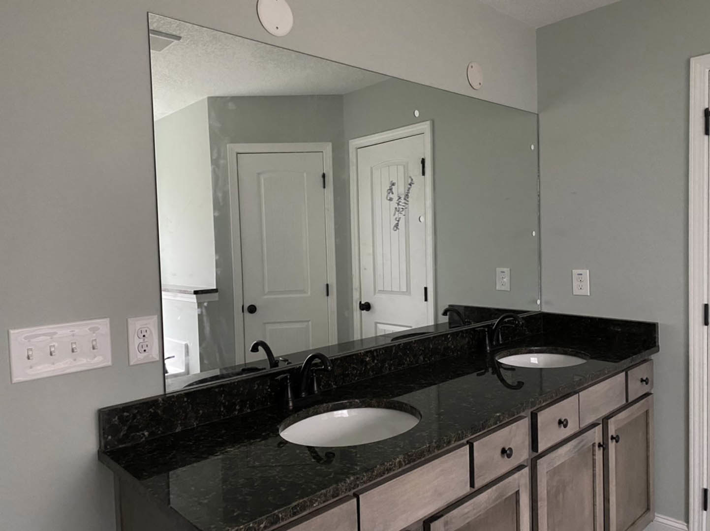 Bathroom with double vanity featuring white oval sinks set in black marble countertop, large wall mirror above, white cabinetry with black handles, four-switch white light plate