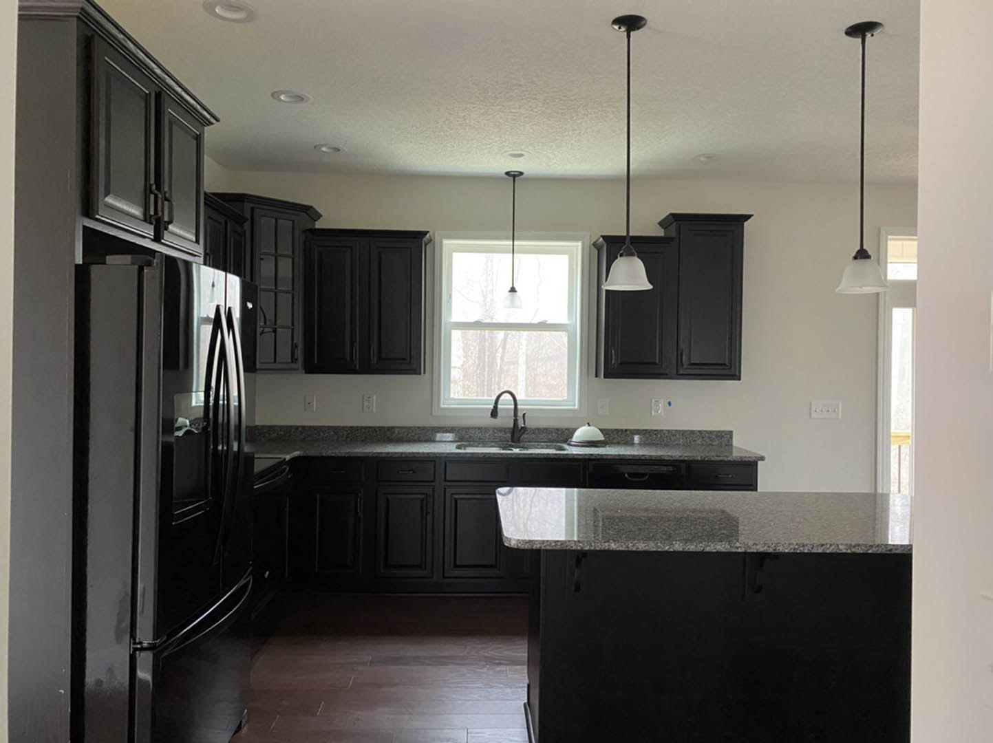 Black kitchen cabinets and refrigerator, black countertop with matching shelf, tile backsplash, stainless steel sink and faucet, ceiling light fixture, white walls.