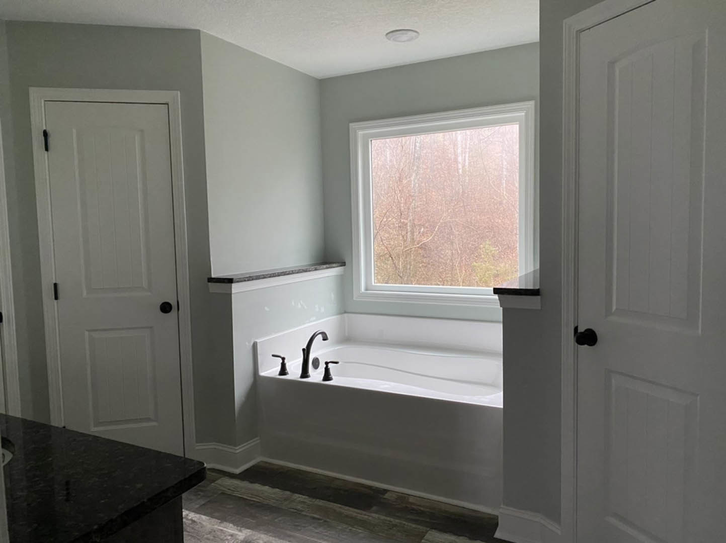 Bathroom with freestanding white bathtub, chrome faucet and black shower head, large window showing trees outside, black countertop, white door with black hardware, plaster walls