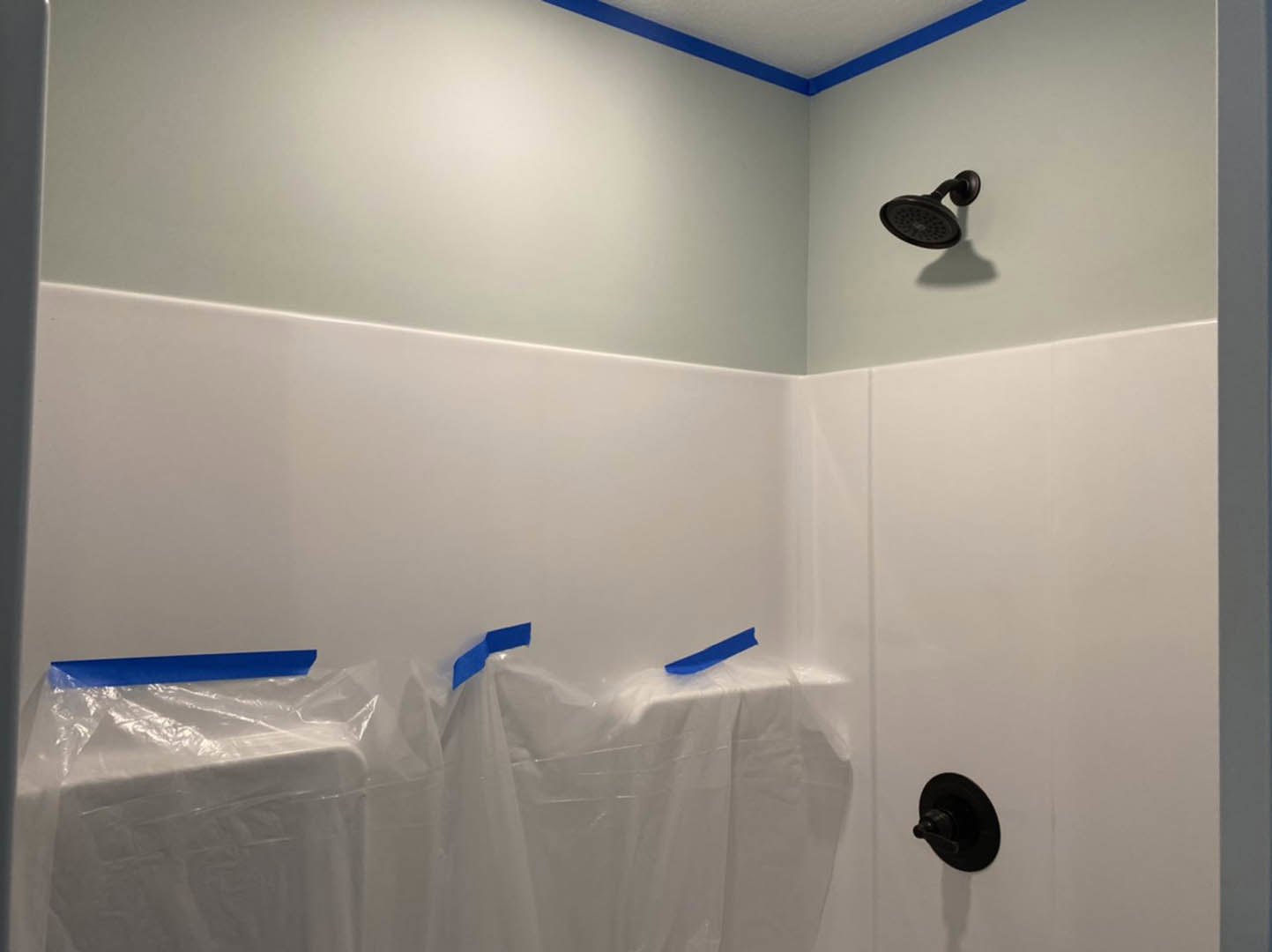 Shower head installed above a wall partially covered with clear plastic sheeting secured by blue tape in a bathroom under construction