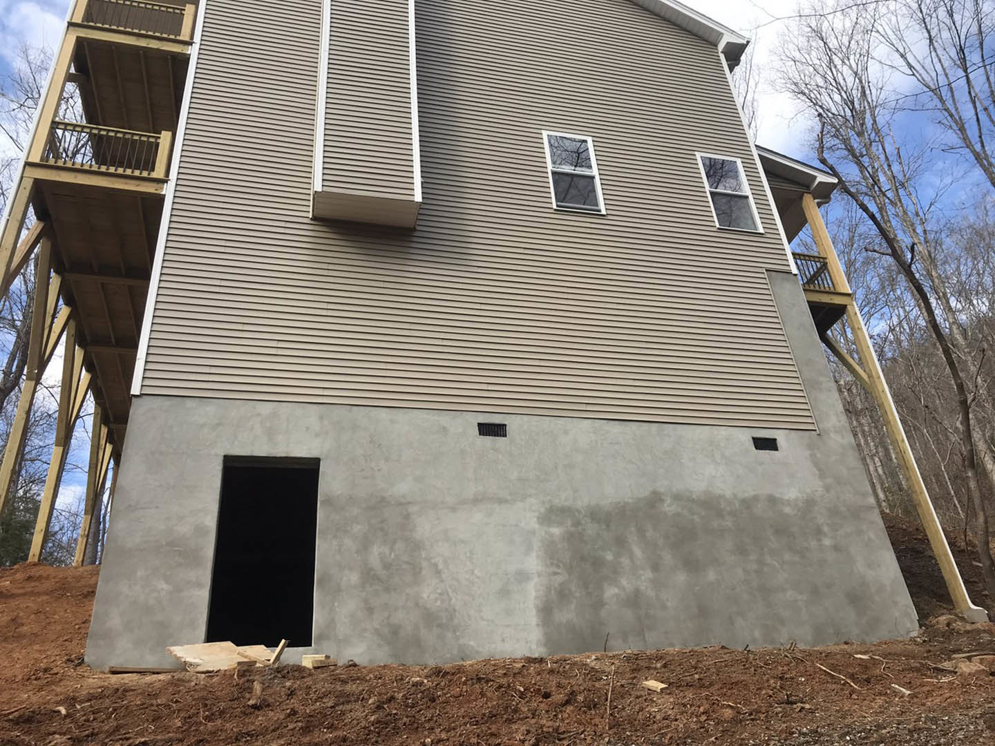 Two-story house under construction featuring a cement wall, balcony with metal railing, large windows, and exposed dirt hill in the foreground