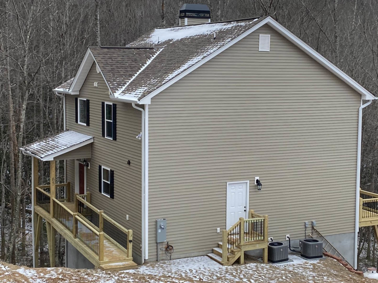 Two-story home with gray siding, white trim, and large windows; wooden deck with metal railing; snow covering roof and ground; white door with black hardware; wooden stairs leading