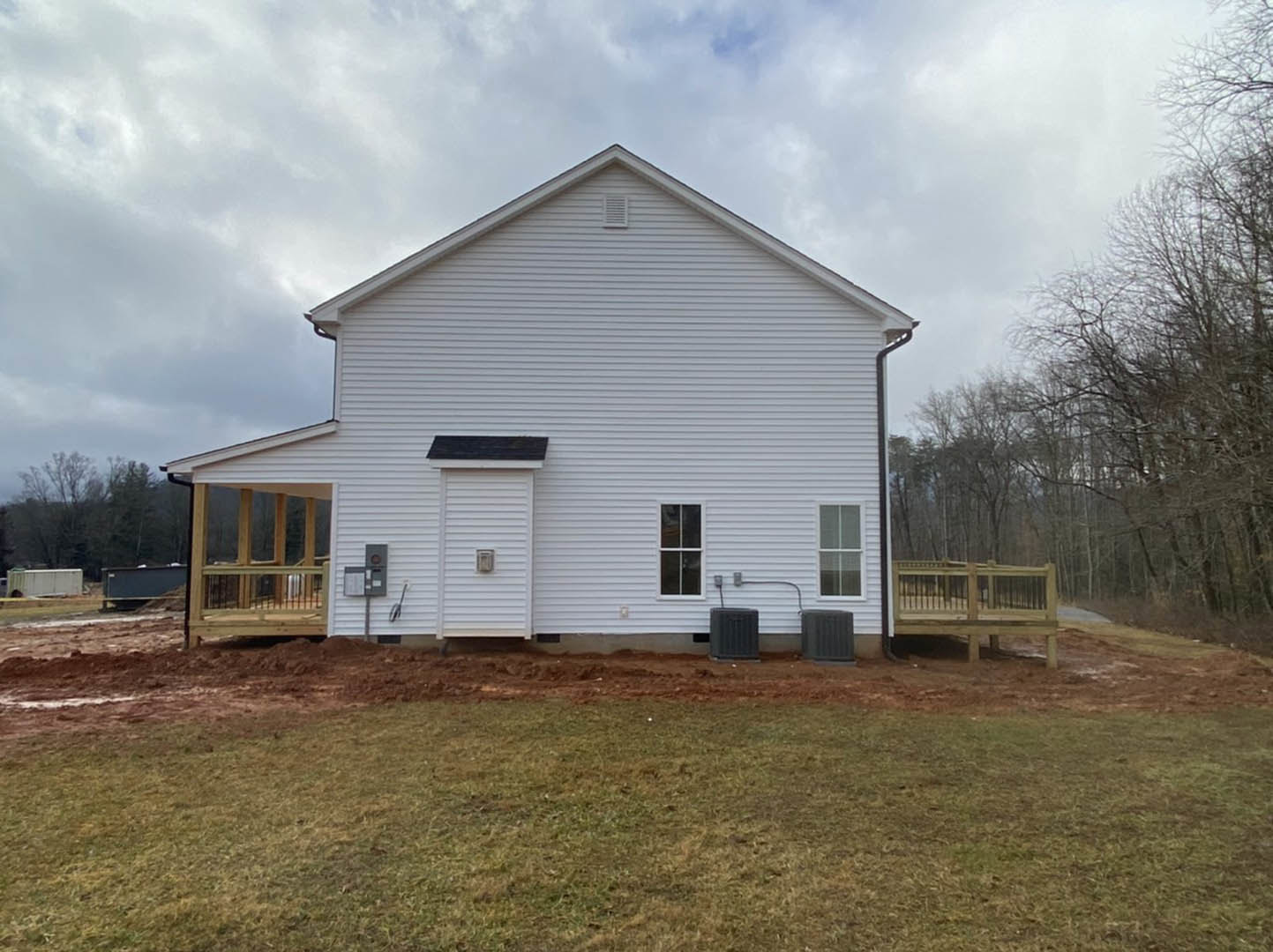 White cottage-style home with open door, wood deck, fenced yard, grassy lawn, and trees under cloudy sky
