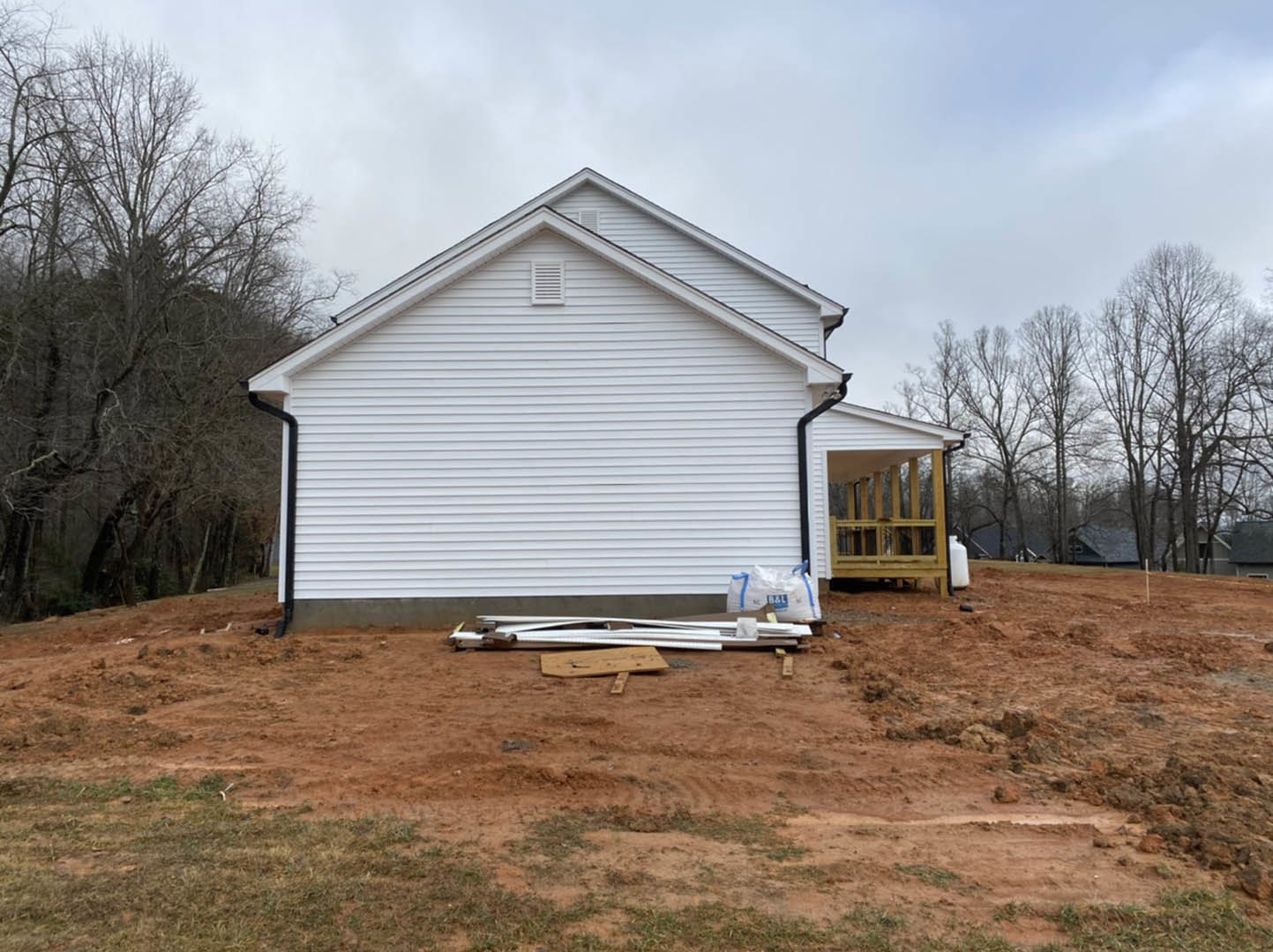 White house under construction with exposed wood framing, pile of lumber on dirt foreground, trees and cloudy sky in background