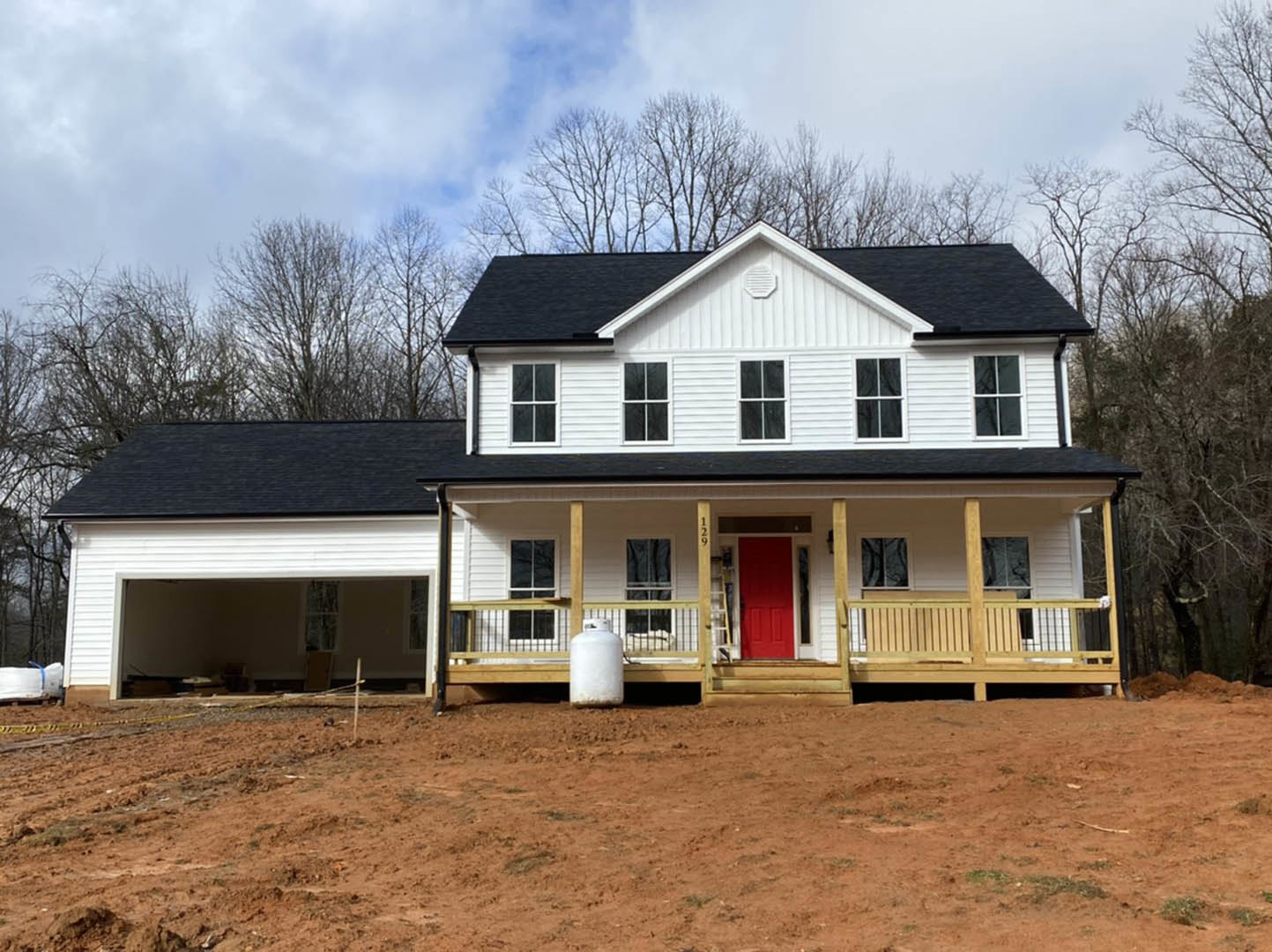 Two-story house under construction with exposed framing, red front door with white trim, covered porch, dirt yard, white cylindrical utility feature, cloudy sky overhead