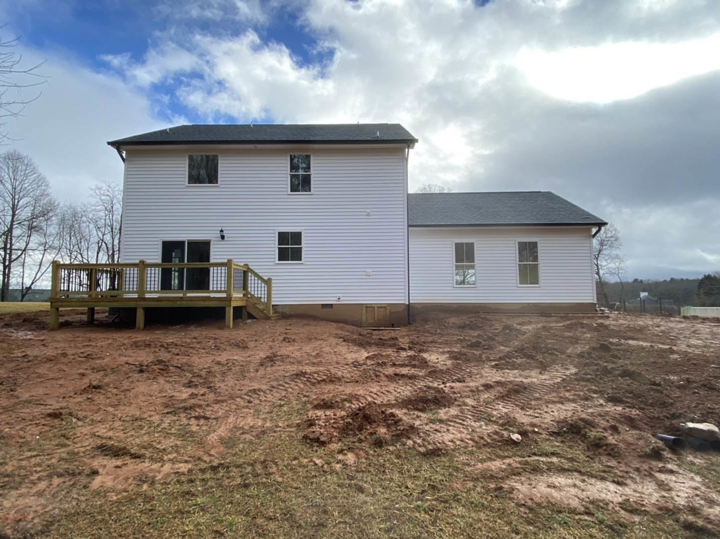 Wood-framed deck with railing attached to partially constructed house, dirt area with tire tracks in foreground, white-framed window, grey siding, blue sky overhead.