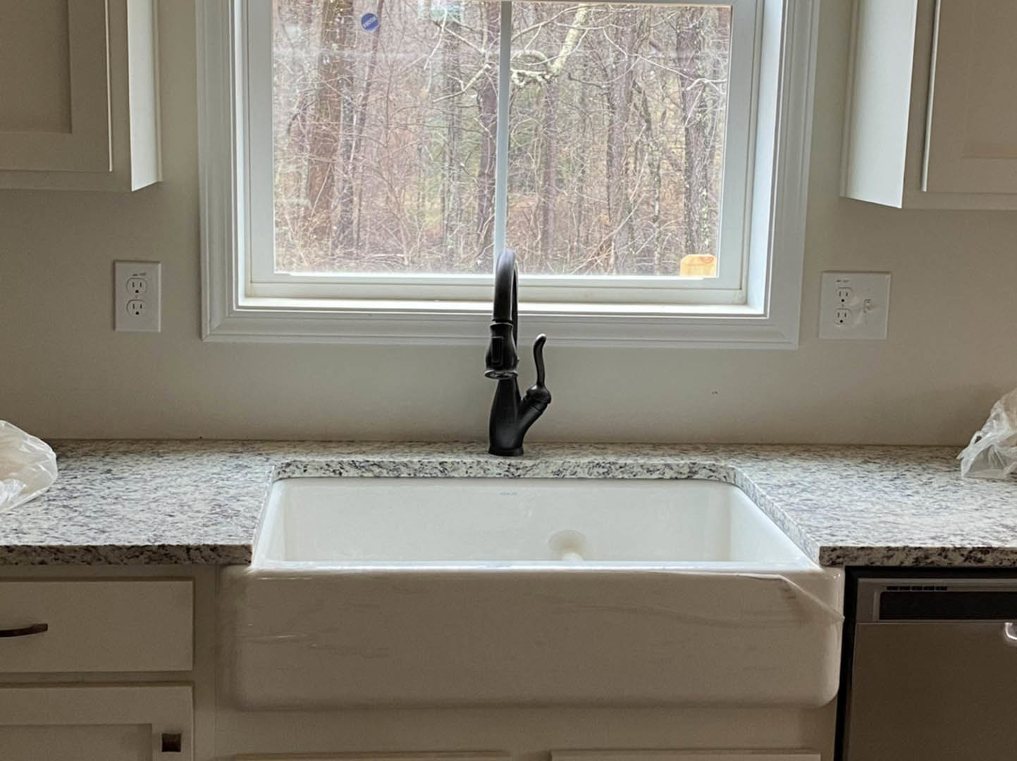 Stainless steel kitchen sink with modern chrome faucet set in quartz countertop, window above reveals leafy trees outside, white cabinetry and electrical outlet visible on